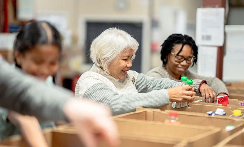 A small group of four volunteers are seen standing at a table full of cardboard boxes as they pack them with non-perishable food items at a local Food Bank.