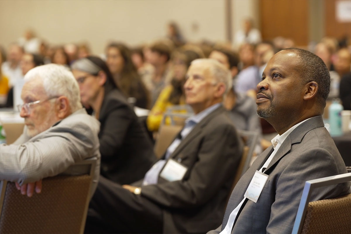 A man in a chair amidst a gathering of conference attendees.