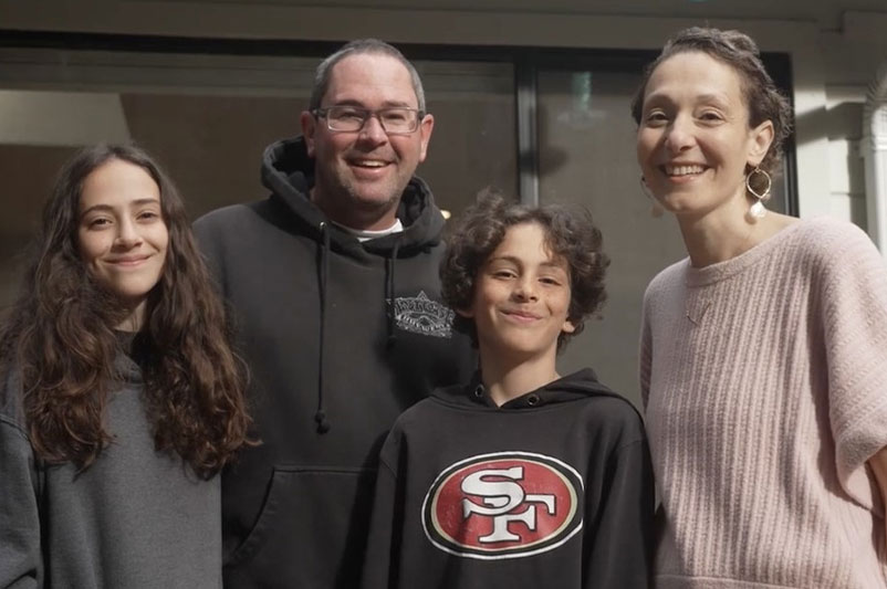 A happy family standing together outside, smiling for a photo on a bright day.